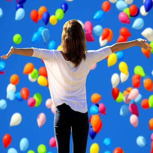A confident woman in a shower of colourful balloons arms out wide