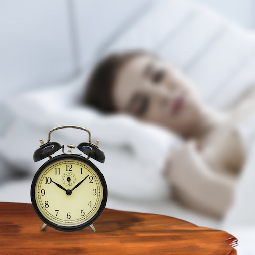 An alarm clock showing 10am and in the background a lady fast asleep in bed